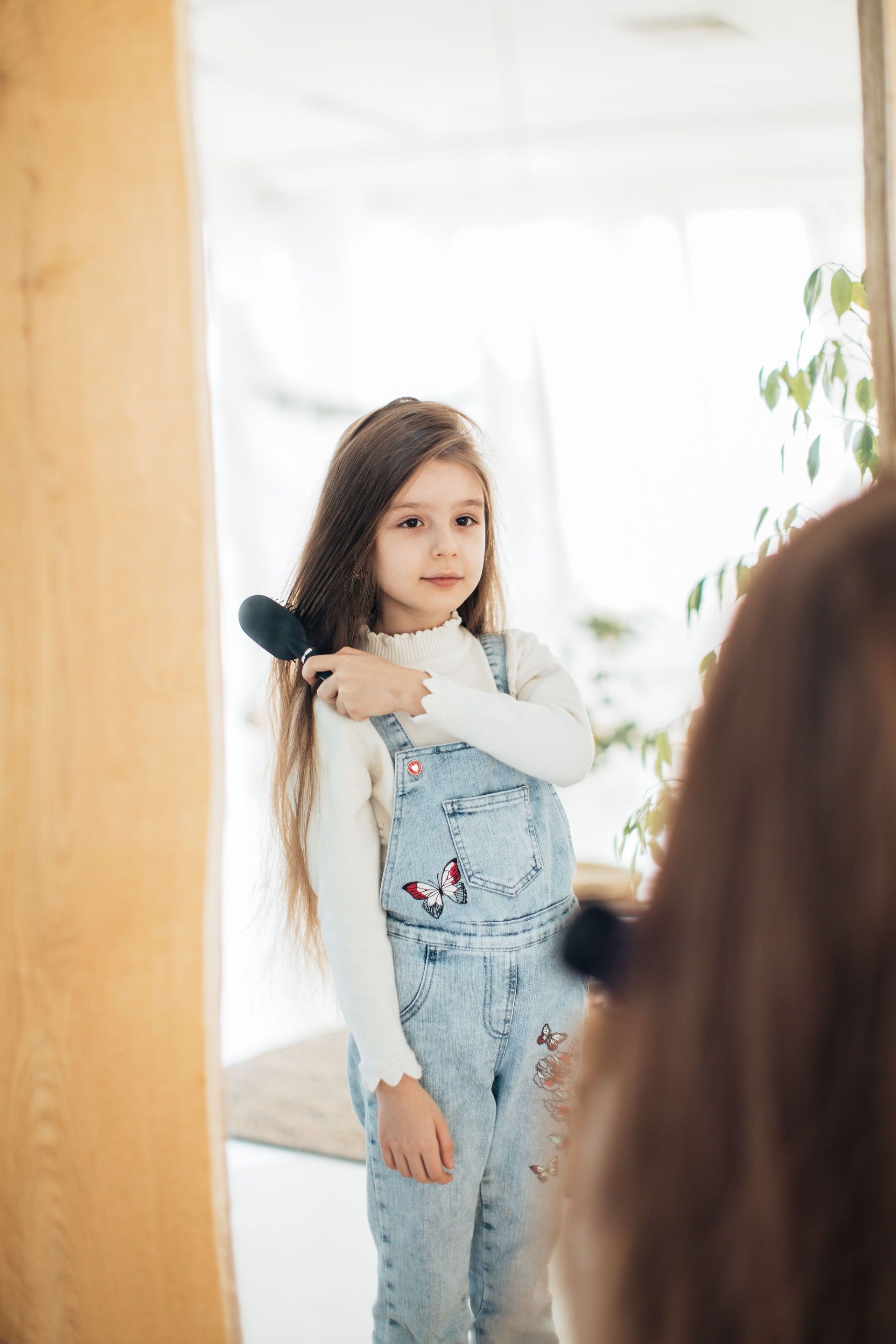 A cute young girl in denim overalls combs her hair while looking in the mirror indoors.