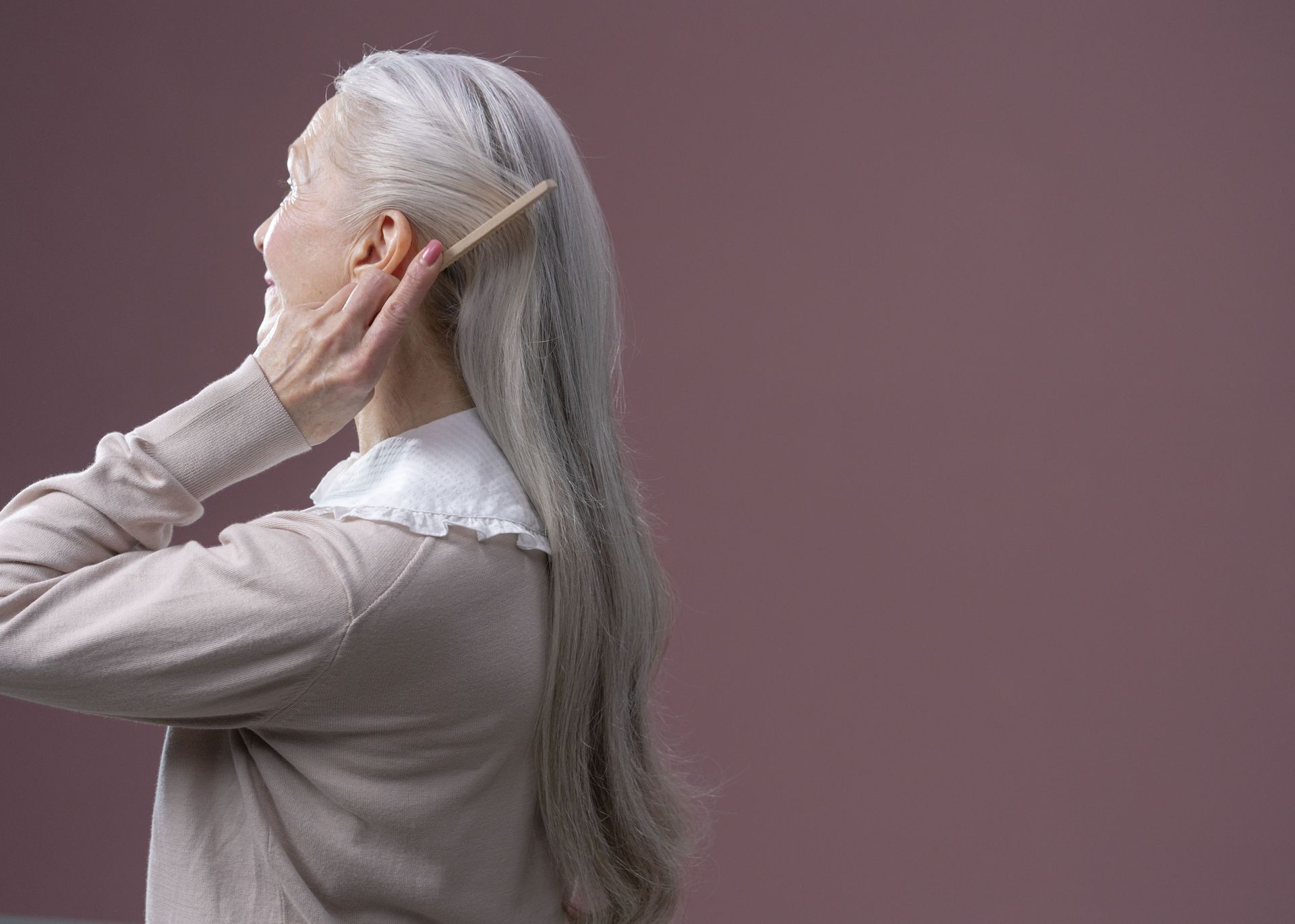 Elderly woman with long grey hair combing it, set against a purple background.