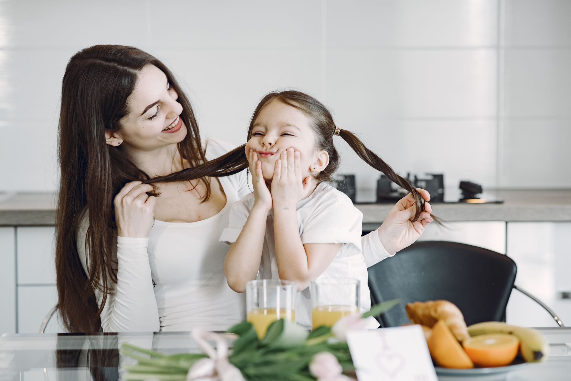 Cheerful mother and daughter in domestic clothes sitting together at table and playing with each other while enjoying breakfast in modern kitchen
