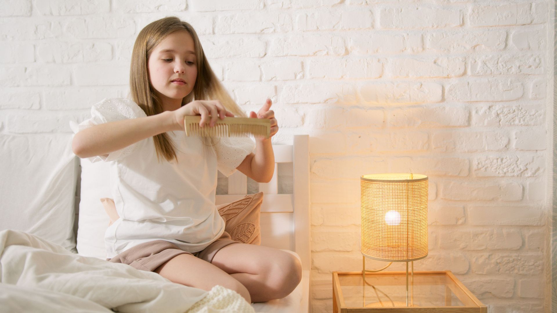 A young girl in a white dress sits on a bed combing her hair beside a warm-lit lamp.