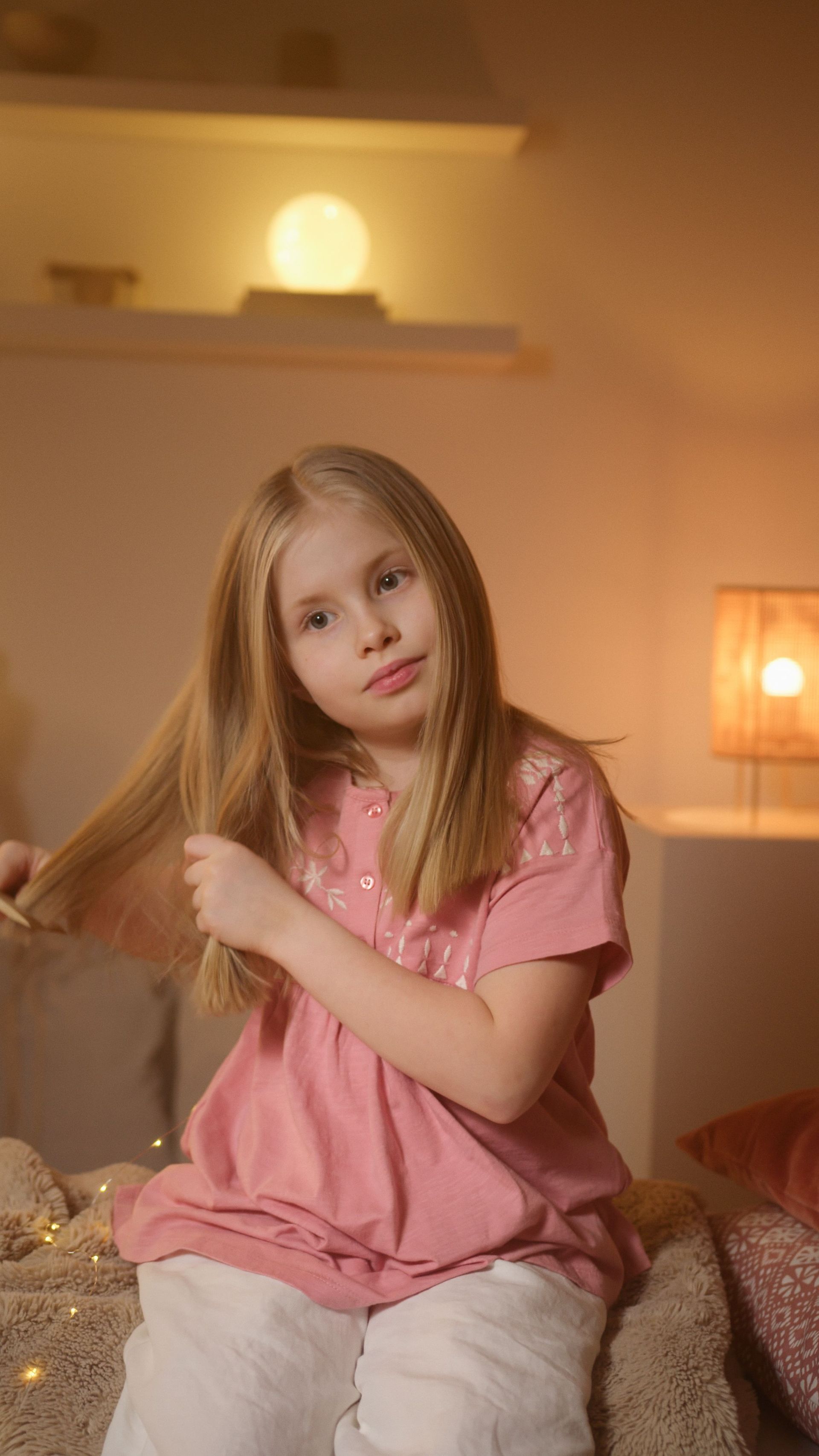 A young girl in a pink shirt gently combs her hair in a warmly lit bedroom, creating a serene and cozy atmosphere.